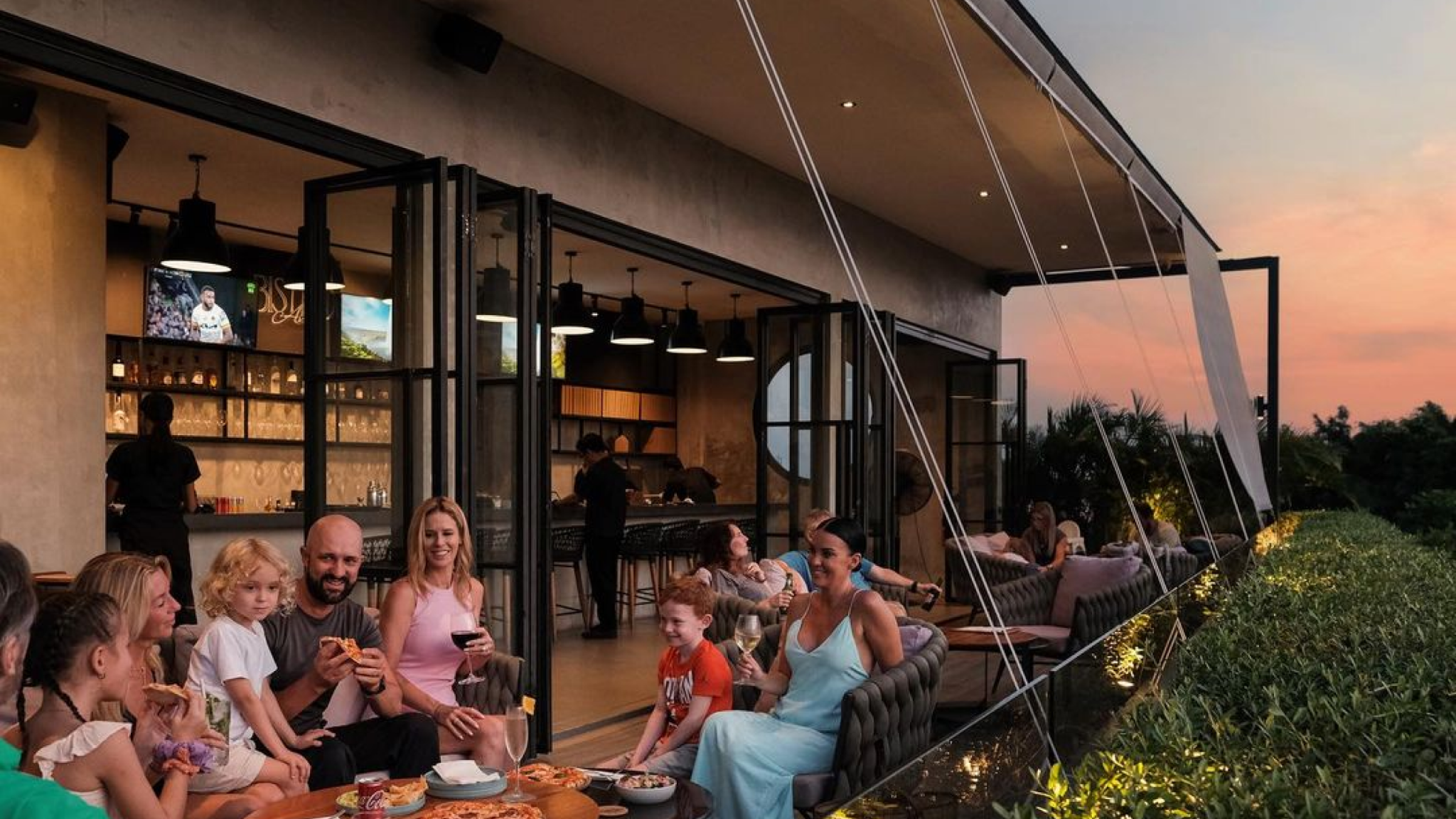 Family and friends enjoying food and drinks at an open air restaurant terrace during sunset.