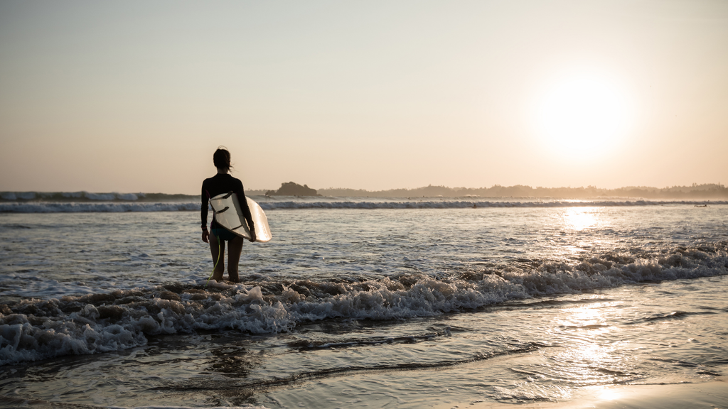 Surfer carrying a surfboard into the ocean during a peaceful sunset.