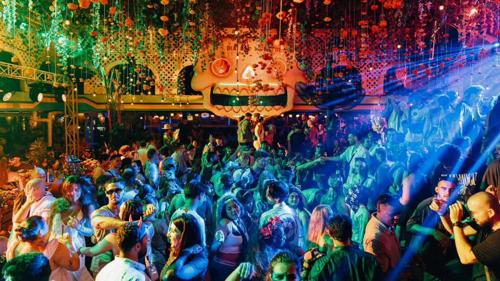 Crowded nightclub filled with people dancing under colorful lights and vibrant tropical decorations.